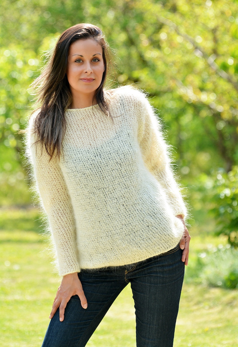 May include: A white fuzzy mohair sweater with a round neckline. The sweater is loose fitting and has long sleeves. The model is wearing the sweater with blue jeans.