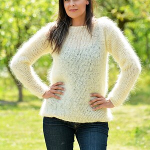 May include: A white fuzzy mohair sweater with long sleeves and a round neckline. The sweater is worn by a person with long brown hair and is photographed outdoors in a green grassy field.