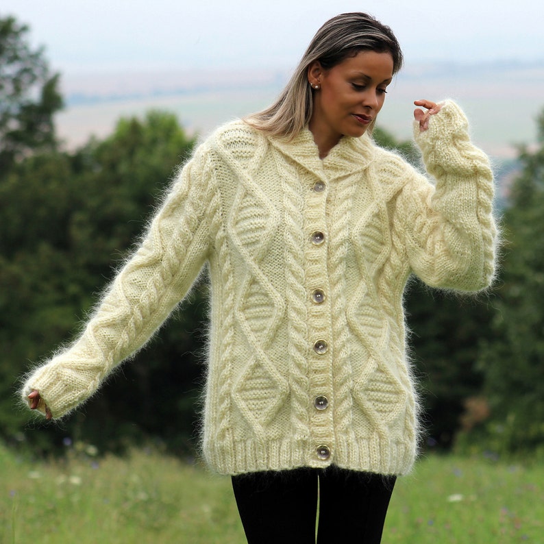 May include: A cream-colored fuzzy cardigan with a cable knit pattern and button closure. The cardigan has a collar and long sleeves.