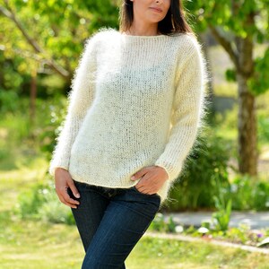 May include: A white fuzzy mohair sweater with a round neckline and long sleeves. The sweater is worn by a person with long dark hair and is photographed outdoors in a green grassy area.
