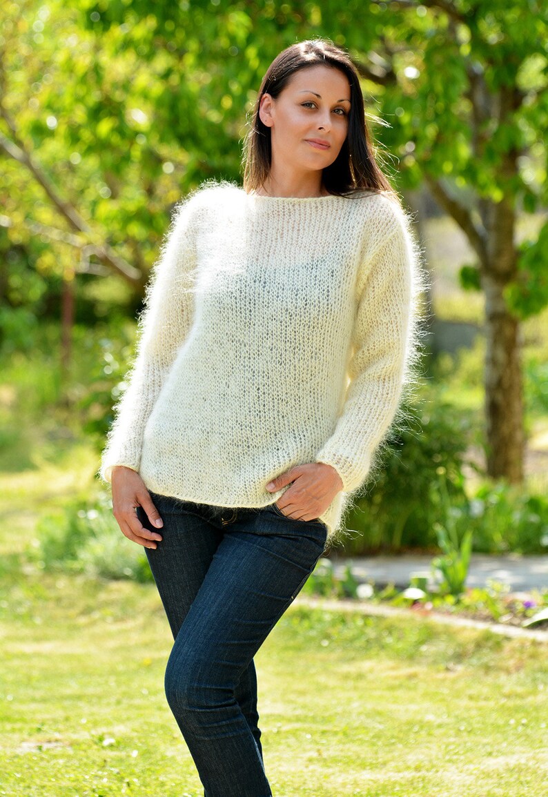 May include: A white fuzzy mohair sweater with a round neckline and long sleeves. The sweater is worn by a person with long dark hair and is photographed outdoors in a green grassy area.