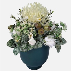 May include: An artificial flower arrangement in a teal ceramic pot. The arrangement features a large white protea flower, white roses, and green foliage.