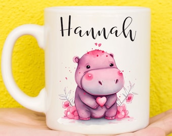 May include: White ceramic mug with a pink hippopotamus illustration holding a heart. The mug has the name "Hannah" written in black cursive on the front.