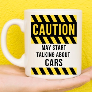 May include: White ceramic mug with the text "CAUTION MAY START TALKING ABOUT CARS" in black font. The text is framed by yellow and black hazard stripes. The mug has a curved handle and is held against a yellow background.