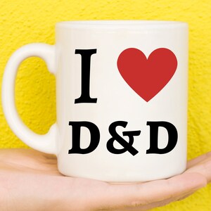 Peut inclure: Une tasse en céramique blanche avec un cœur rouge et le texte "I love D&D" imprimé dessus.