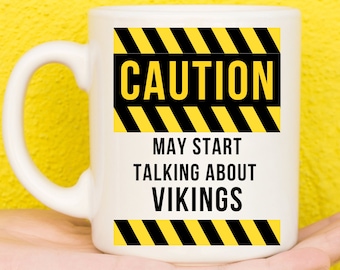Puede incluir: Taza de cerámica blanca con una señal de precaución amarilla y negra que dice "PRECAUCIÓN PUEDE COMENZAR A HABLAR DE VIKINGOS".