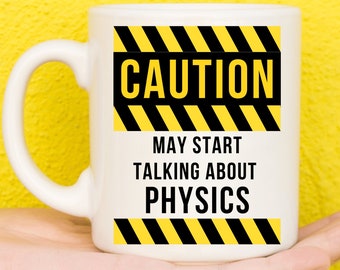 May include: White coffee mug with a black and yellow caution sign that reads "CAUTION MAY START TALKING ABOUT PHYSICS".