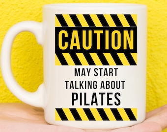 Op de afbeelding: Witte keramische mok met een zwart-geel waarschuwingsbord met de tekst "LET OP KAN BEGINNEN OVER PILATES TE PRATEN".