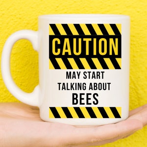 May include: White ceramic mug with a black and yellow caution sign that reads "CAUTION MAY START TALKING ABOUT BEES".