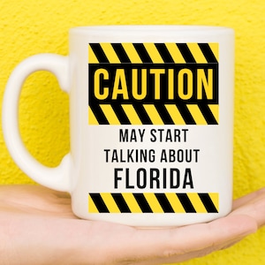 May include: White ceramic mug with the text "CAUTION MAY START TALKING ABOUT FLORIDA" in black letters. The mug has yellow and black hazard stripes above and below the text. The mug is held in a hand against a yellow background.