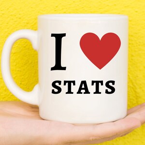 May include: White ceramic mug with the text "I ♥ STATS" in black and a red heart symbol. The mug has a curved handle and is held in a person's hand against a yellow background. The mug is a standard size.