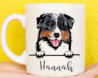 Peut inclure: Une tasse en céramique blanche avec une illustration de dessin animé d'un berger australien regardant par-dessus le bord. Le chien a un pelage noir, brun et blanc et regarde directement le spectateur. La tasse porte le nom "Hannah" écrit en cursive noire sur le devant.