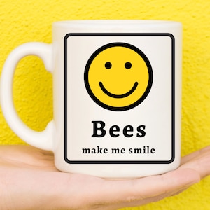 May include: White ceramic mug with a black border around a yellow smiley face and the text "Bees make me smile".