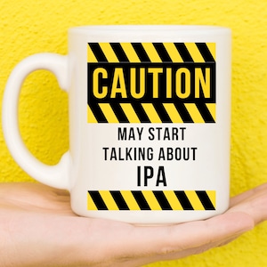 May include: White ceramic mug with a black and yellow caution sign that reads "CAUTION MAY START TALKING ABOUT IPA".