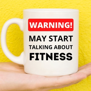 Może przedstawiać: Biały kubek ceramiczny z czerwono-czarną tabliczką ostrzegawczą z napisem "UWAGA! MOŻE ZACZĄĆ ROZMOWĘ O FITNESS".