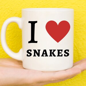 Puede incluir: Taza de café de cerámica blanca con un corazón rojo y el texto "Me encantan las serpientes" en negro.