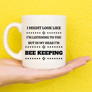May include: A white ceramic mug with a black text design. The text reads "I MIGHT LOOK LIKE I'M LISTENING TO YOU BUT IN MY HEAD I'M BEE KEEPING". The mug has a curved handle and is held against a yellow background.