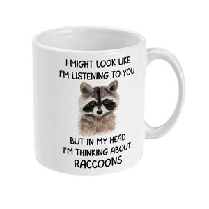May include: White ceramic mug with a black and white illustration of a raccoon. The mug has the text "I might look like I'm listening to you, but in my head I'm thinking about raccoons."