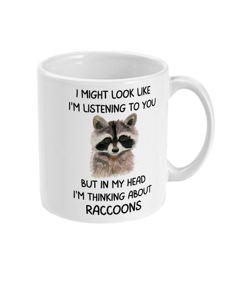 May include: White ceramic mug with a black and white illustration of a raccoon. The mug has the text "I might look like I'm listening to you, but in my head I'm thinking about raccoons."