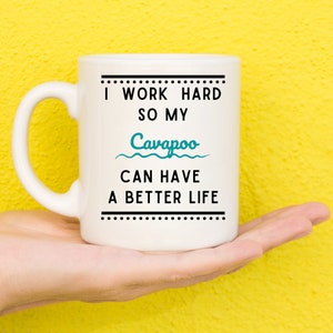 May include: White ceramic mug with the text "I WORK HARD SO MY Cavapoo CAN HAVE A BETTER LIFE" in black and teal. The mug has a curved handle and is held in a hand against a yellow background.