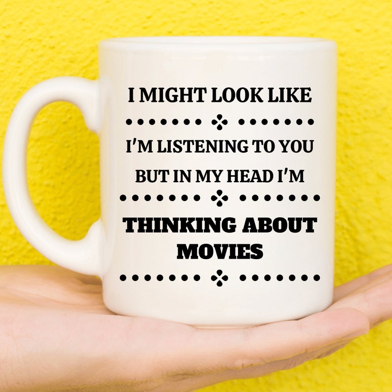 May include: White ceramic mug with the text "I MIGHT LOOK LIKE I'M LISTENING TO YOU BUT IN MY HEAD I'M THINKING ABOUT MOVIES" in black. The mug has a curved handle and is held against a yellow background.