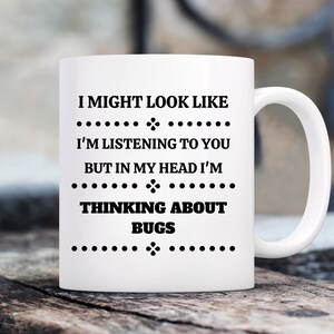 May include: White ceramic coffee mug with black text that reads "I might look like I'm listening to you but in my head I'm thinking about bugs".