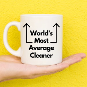 Peut inclure: Une tasse à café en céramique blanche avec du texte noir qui dit "World's Most Average Cleaner" avec deux flèches noires pointant vers le haut.