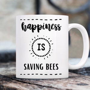 May include: White ceramic coffee mug with black text that reads "happiness is saving bees".