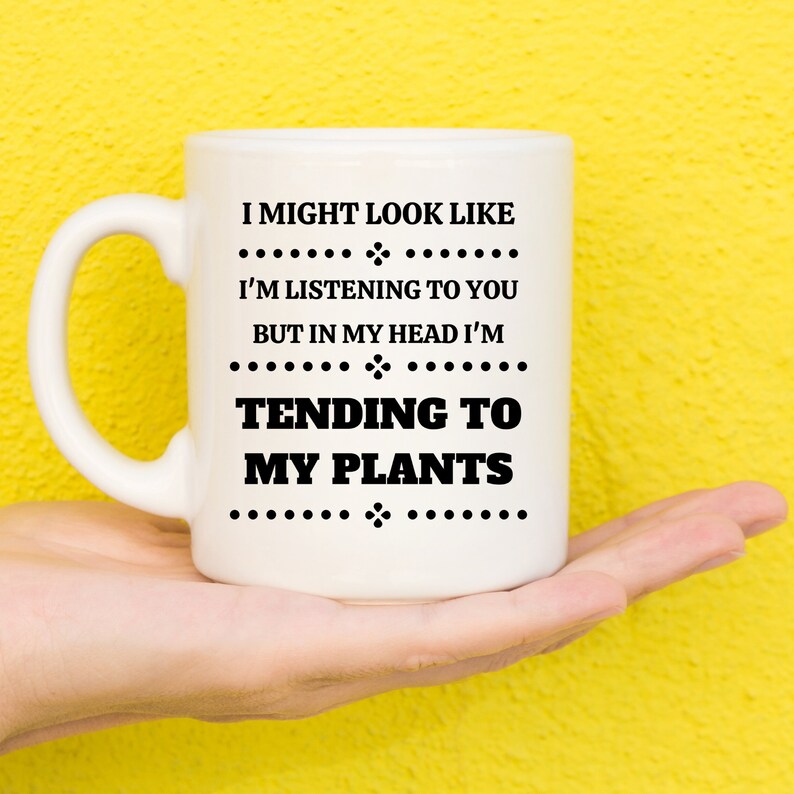 May include: White ceramic mug with the text "I MIGHT LOOK LIKE I'M LISTENING TO YOU BUT IN MY HEAD I'M TENDING TO MY PLANTS" in black. The mug is held against a yellow background.