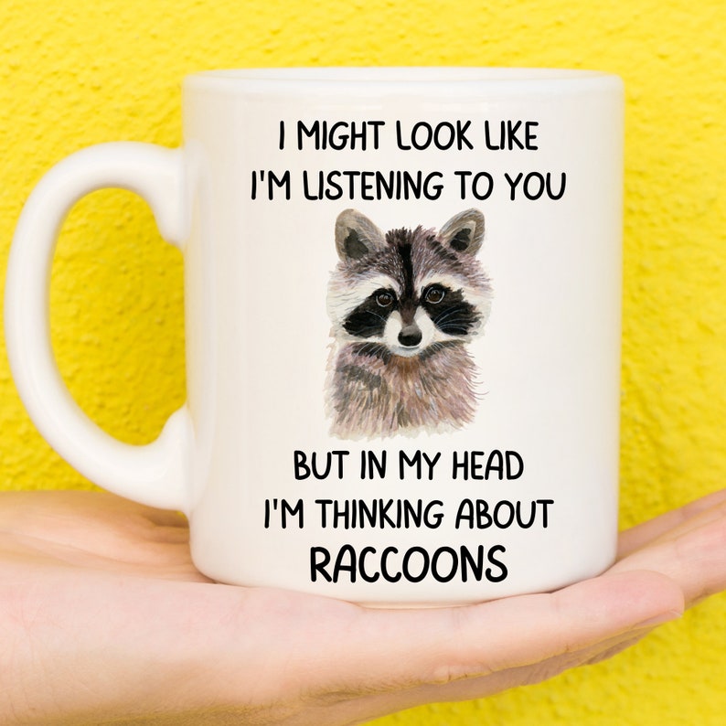 May include: White ceramic mug with a black and gray raccoon illustration. The mug has the text "I might look like I'm listening to you, but in my head I'm thinking about raccoons."