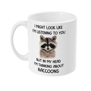 May include: White ceramic coffee mug with a black and white illustration of a raccoon. The mug has the text "I might look like I'm listening to you, but in my head I'm thinking about raccoons."