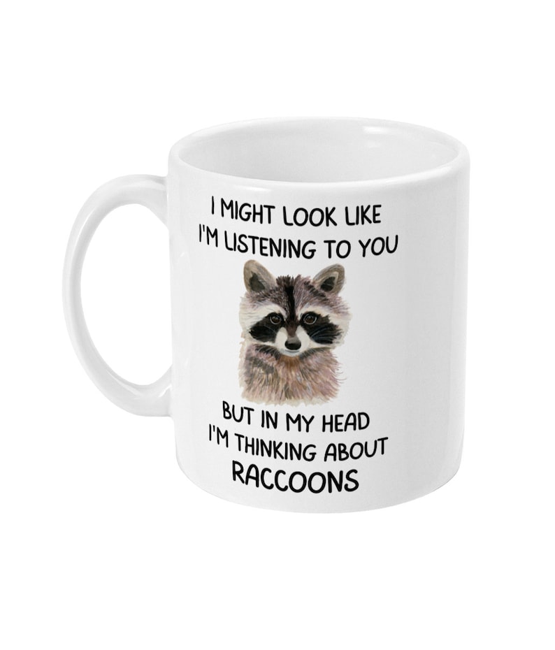 May include: White ceramic coffee mug with a black and white illustration of a raccoon. The mug has the text "I might look like I'm listening to you, but in my head I'm thinking about raccoons."