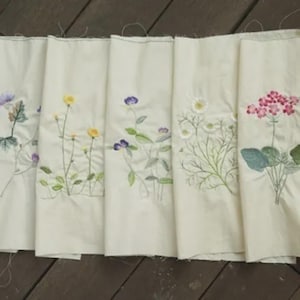 Peut inclure: Plusieurs panneaux de tissu blanc cassé, chacun brodé d'un motif floral différent. Les fleurs comprennent du chardon violet, des boutons d'or jaunes, des gloires du matin violettes, des marguerites blanches et des géraniums roses. La broderie est détaillée et colorée.