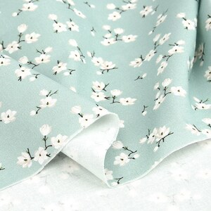 May include: A light sage green fabric featuring a repeating pattern of small white blossoms with dark brown stems. The textile is folded to reveal a textured weave and a lighter, off-white underside, showcasing the floral print across the surface.