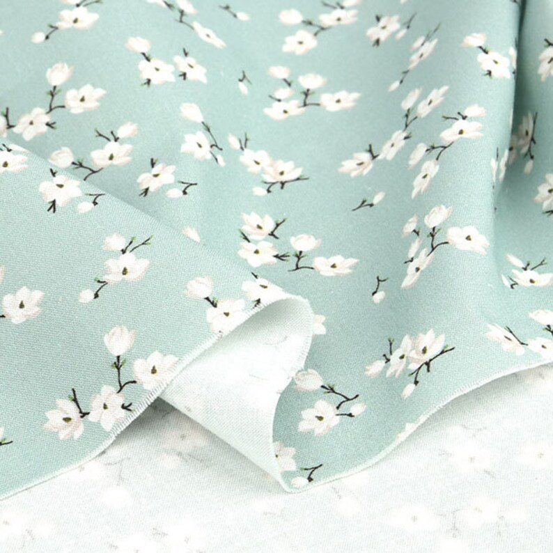 May include: A light sage green fabric featuring a repeating pattern of small white blossoms with dark brown stems. The textile is folded to reveal a textured weave and a lighter, off-white underside, showcasing the floral print across the surface.