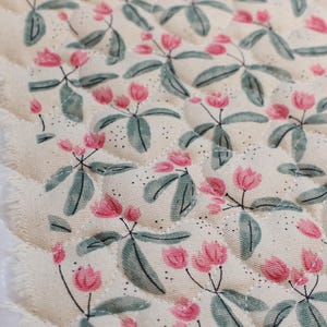Pu&ograve; includere: Un pezzo di tessuto trapuntato con uno sfondo color crema e un motivo ripetuto di piccoli fiori rosa e foglie verde salvia. Il tessuto presenta una trama cucita a rombi e un bordo grezzo e sfilacciato sul lato sinistro del materiale.