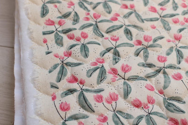 Pu&ograve; includere: Un pezzo di tessuto trapuntato con uno sfondo color crema e un motivo ripetuto di piccoli fiori rosa e foglie verde salvia. Il tessuto presenta una trama cucita a rombi e un bordo grezzo e sfilacciato sul lato sinistro del materiale.