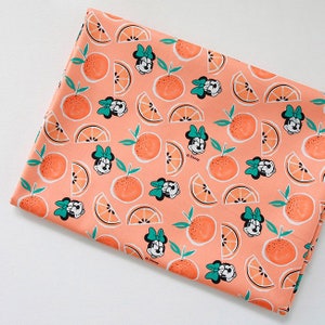 Puede incluir: Tela de color melocot&oacute;n con un patr&oacute;n repetido de rodajas de naranja e im&aacute;genes de dibujos animados de Minnie Mouse con un lazo verde. La tela tiene el texto "Disney" impreso.