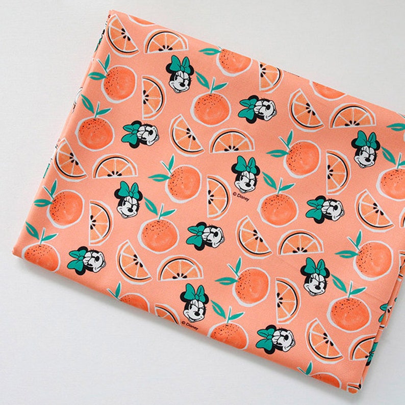 Puede incluir: Tela de color melocot&oacute;n con un patr&oacute;n repetido de rodajas de naranja e im&aacute;genes de dibujos animados de Minnie Mouse con un lazo verde. La tela tiene el texto "Disney" impreso.