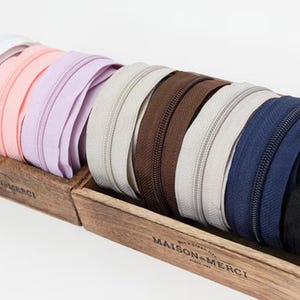 May include: A collection of colorful zippers in a wooden box. The zippers are in various colors, including white, pink, lavender, beige, brown, navy blue, and black. The box has the text "MAISON MERCI" printed on the front.