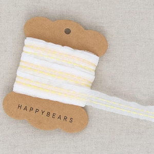 May include: White ribbon with yellow accents, spooled on a brown cardboard tag. The tag reads "HAPPYBEARS" in black text. The ribbon is partially unraveled, showing its texture and design. The background is a neutral fabric.