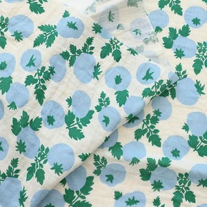 May include: A piece of off-white fabric featuring a repeating pattern of light blue circular shapes and green leafy stems. The textile displays a consistent print of stylized botanical elements distributed across the surface in a balanced layout.