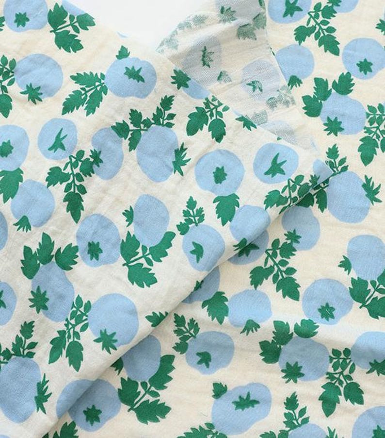 May include: A piece of off-white fabric featuring a repeating pattern of light blue circular shapes and green leafy stems. The textile displays a consistent print of stylized botanical elements distributed across the surface in a balanced layout.