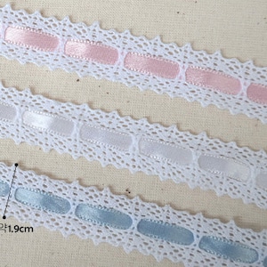 May include: Three white lace trims with different coloured satin ribbon accents. The top trim has pink ribbon, the middle trim has white ribbon, and the bottom trim has light blue ribbon. Each trim is approximately 1.9 cm wide.