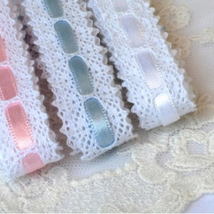 May include: Three white lace trims with different coloured satin ribbon accents. The trims feature a delicate lace pattern and are available in pink, blue, and white.