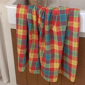 May include: A rectangular fabric towel featuring a vibrant plaid pattern with intersecting lines in shades of red, yellow, blue, and green. The textile is draped over the edge of a white basin, displaying a consistent grid-like design throughout.