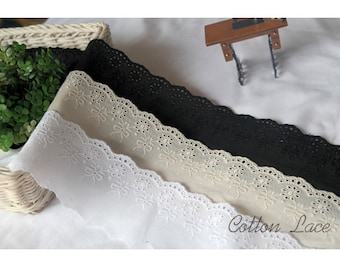 Peut inclure: Trois rouleaux de dentelle en coton de couleur blanche, beige et noire. La dentelle a un bord festonné et un motif floral délicat. Le texte "Cotton Lace" est visible dans le coin inférieur droit de l'image.