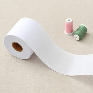May include: A roll of white fabric with a visible texture, unrolled to reveal its width. Two spools of thread, one pink and one green, are in the background. The fabric roll has a brown cardboard core.
