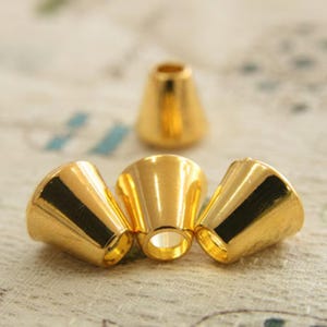 May include: Close-up of several gold-colored cone-shaped jewelry findings. The findings have a polished, reflective surface and a flared opening. One finding is in the background, while three are in the foreground, slightly overlapping.