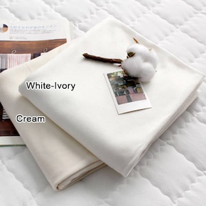 May include: Two folded fabric samples, one cream and one white-ivory, are displayed on a quilted surface. A sprig of cotton and a small photo card rest on the white-ivory fabric. The text "White-Ivory" and "Cream" are visible.